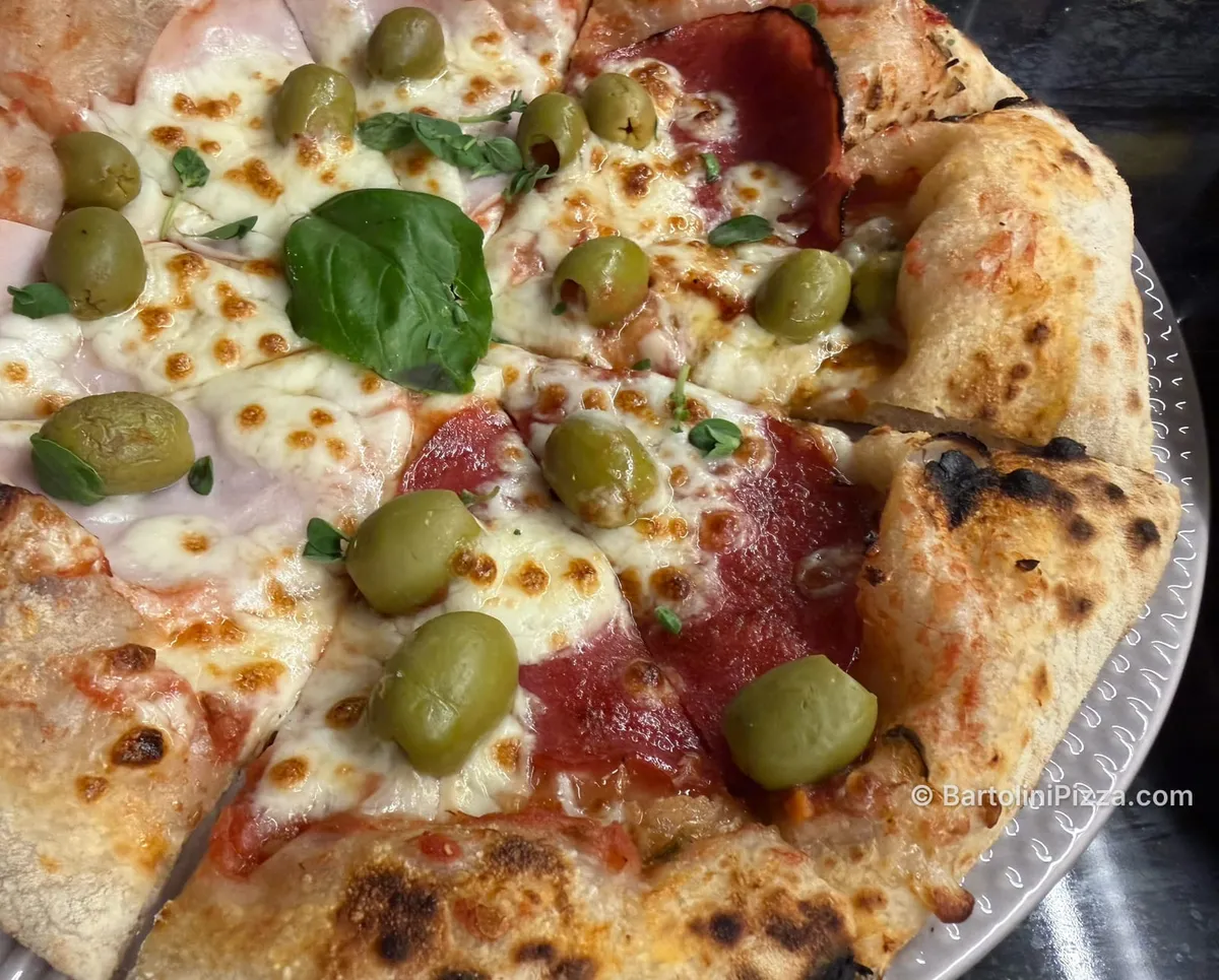 Close-up of a sliced homemade pizza with green olives, ham, salami and a basil leaf.